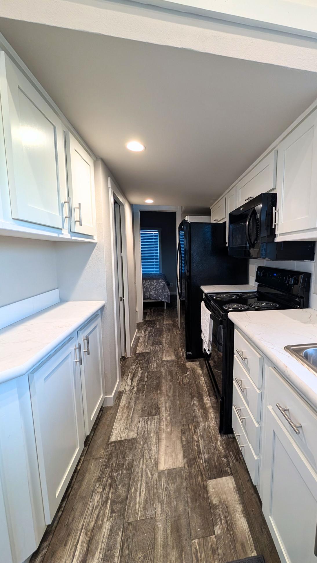 Narrow galley kitchen with white cabinets, black appliances, wood-look flooring, and a view toward a bedroom at the end of the hallway.