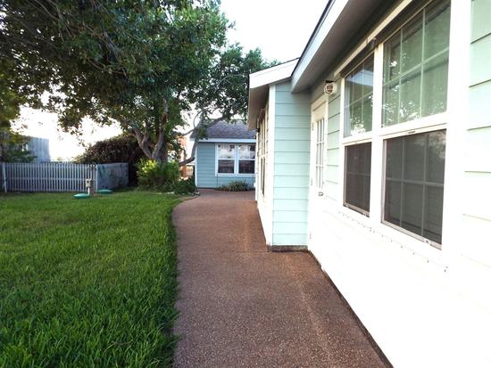 Side yard of a coastal home with a concrete walkway, green grass, trees for shade, and light blue exterior siding.