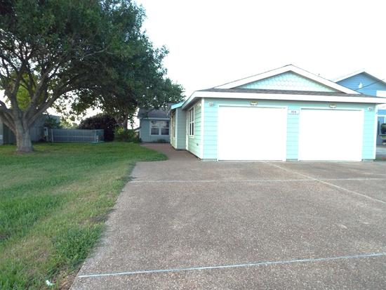 Concrete driveway with multiple parking spaces in front of a light blue garage, with a grassy yard and trees nearby.