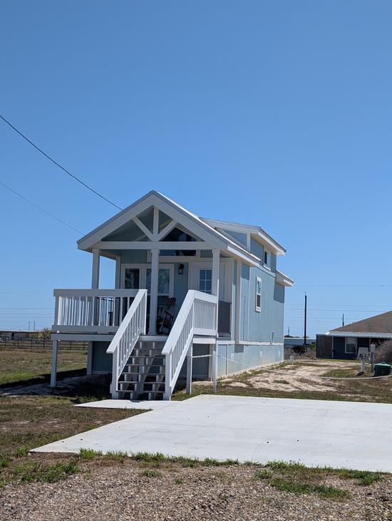 Light blue elevated coastal cottage with white trim, front stairs leading to a covered porch, and a large concrete driveway under a clear blue sky