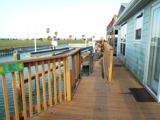 Wooden deck along the water with railing, attached to a light blue coastal home, leading down to a dock with boats and canal views.