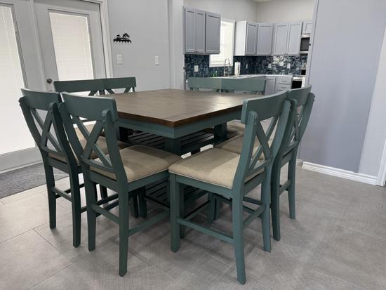 Coastal-style dining area with seating for six, blue-green chairs, wood dining table, and open view into a modern kitchen with beach-inspired finishes in a bright vacation rental.