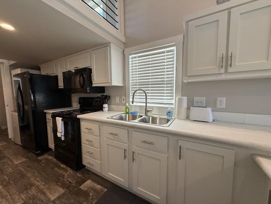 Bright vacation rental kitchen featuring white shaker cabinets, black appliances, double stainless sink, high-arc faucet, wood-look flooring, and tall ceilings with loft overlook in a clean coastal-style interior.