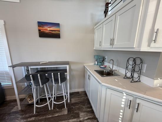 Coastal-style kitchenette area with white cabinets, small sink, breakfast bar seating for two, wood-look flooring, and beach sunset wall art in a bright vacation rental interior.