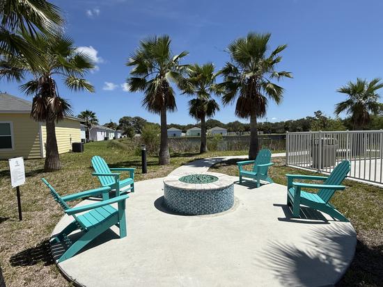 Outdoor community fire pit area with turquoise Adirondack chairs, palm trees, and sunny coastal landscaping near vacation cottages under a bright blue sky.