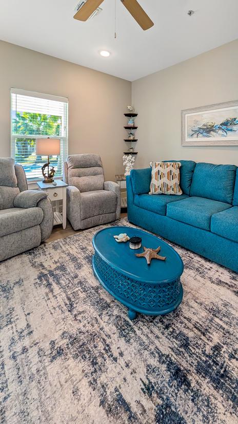 Living room with a blue sofa, two recliners, oval coffee table, area rug, side table with lamp, and coastal wall art.