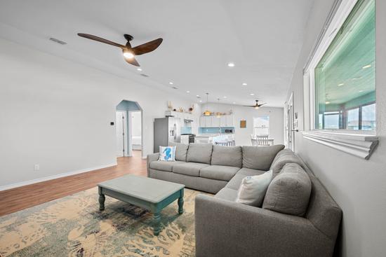 Open-concept living room with a large gray sectional, coastal coffee table, patterned area rug, and kitchen beyond.