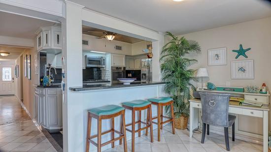 Kitchen with stainless steel appliances, granite breakfast bar with three teal bar stools, and a small white desk with a gray chair.