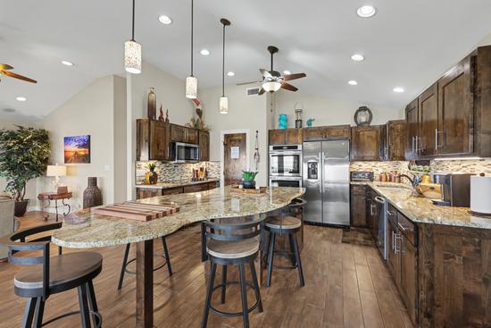 Kitchen with curved granite island and four bar stools, dark wood cabinets, stainless refrigerator, double wall oven, microwave, and pendant lights.