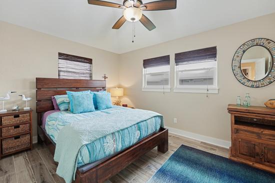 Bedroom with queen bed in a dark wood frame, turquoise bedding, wicker-drawer nightstand, wood dresser, round mosaic mirror, and blue rug.