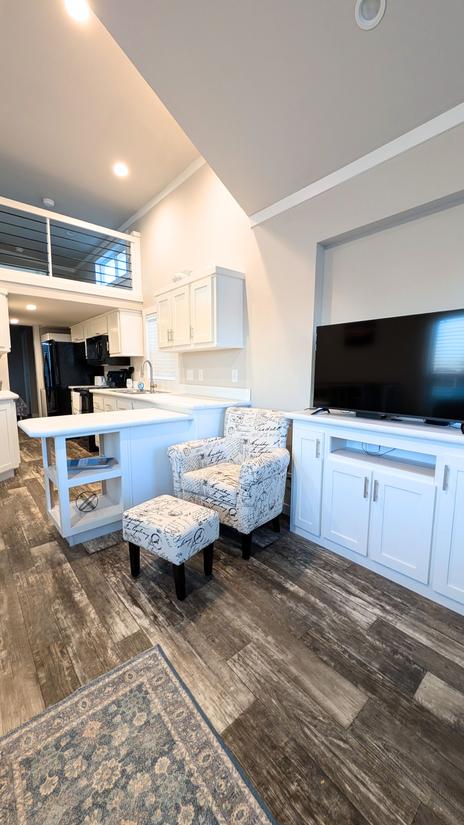 Open living area with a small seating nook, flat-screen TV, white kitchen with breakfast bar, and a loft above, featuring high ceilings and wood-look flooring.